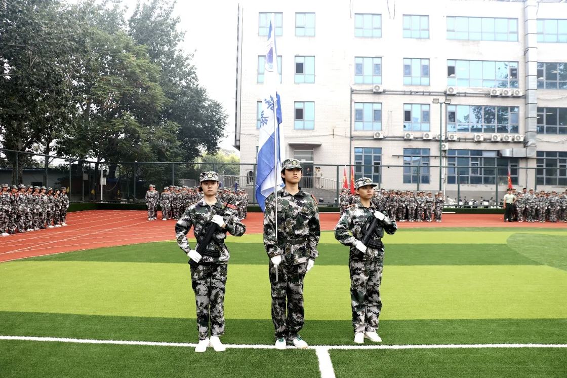 照片：军训结训仪式现场——三名新生组成护旗组合，持系旗与训练用枪在球场肃立，后方各新生方队列队，背景为教学楼（学院官方微信文章配图，2026-09-19 收录）